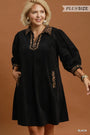 A woman modeling a black plus-size tunic dress with leopard print trim on the collar, neckline, cuffs, and pockets, set against a solid brown studio background.