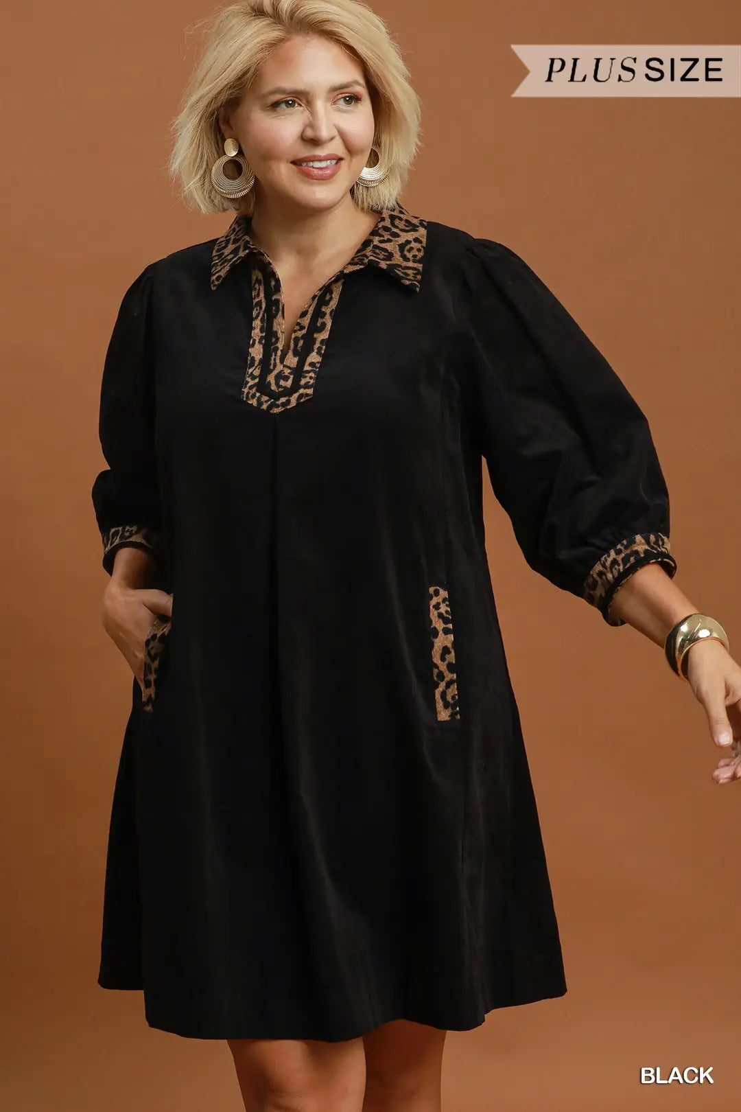 A woman modeling a black plus-size tunic dress with leopard print trim on the collar, neckline, cuffs, and pockets, set against a solid brown studio background.