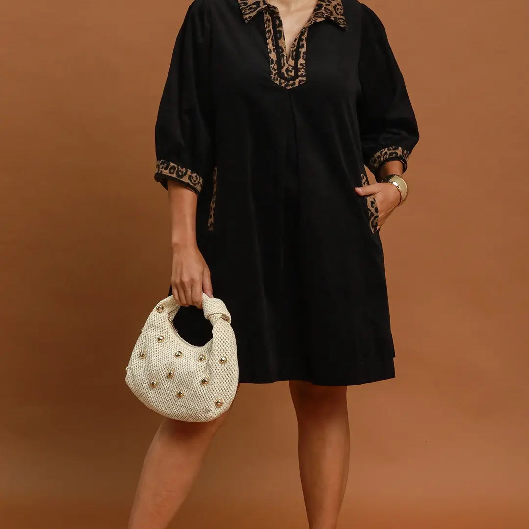 A woman modeling a black short-sleeve tunic dress with gold embroidered trim on the collar and cuffs, holding a white studded handbag against a solid brown studio background. Text overlays read 'PLUS SIZE' and 'BLACK'.