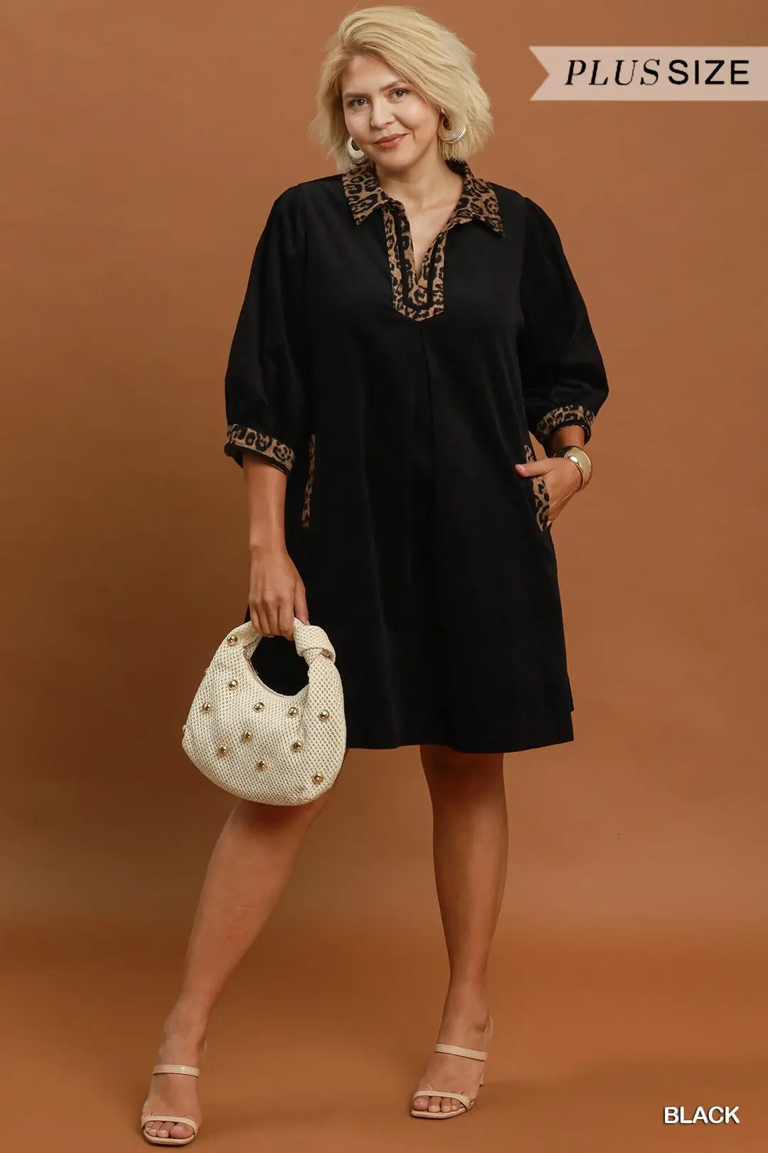 A woman modeling a black short-sleeve tunic dress with gold embroidered trim on the collar and cuffs, holding a white studded handbag against a solid brown studio background. Text overlays read 'PLUS SIZE' and 'BLACK'.
