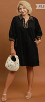 A woman modeling a black short-sleeve tunic dress with gold embroidered trim on the collar and cuffs, holding a white studded handbag against a solid brown studio background. Text overlays read 'PLUS SIZE' and 'BLACK'.