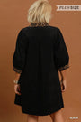 A woman viewed from the back wearing a black short dress or tunic with patterned trim at the collar and sleeve cuffs, against a warm brown studio background. A small 'PLUS SIZE' label appears at the top right, and 'BLACK' is printed at the bottom right.