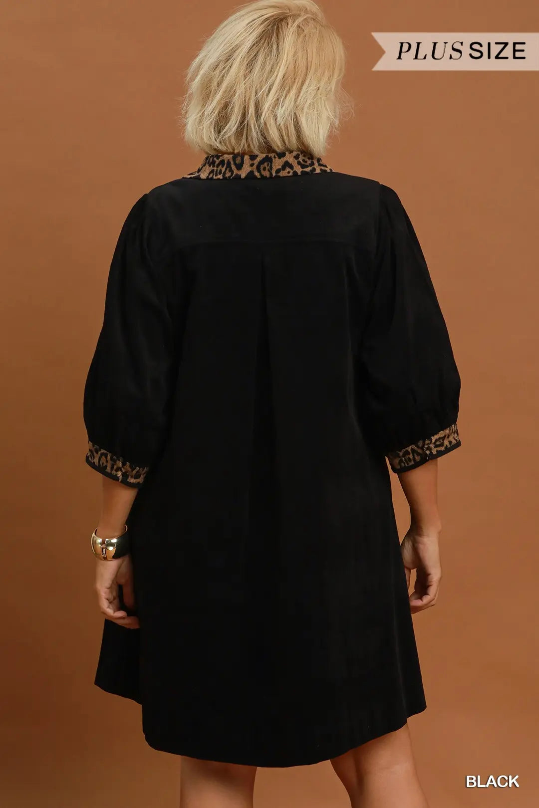 A woman viewed from the back wearing a black short dress or tunic with patterned trim at the collar and sleeve cuffs, against a warm brown studio background. A small 'PLUS SIZE' label appears at the top right, and 'BLACK' is printed at the bottom right.