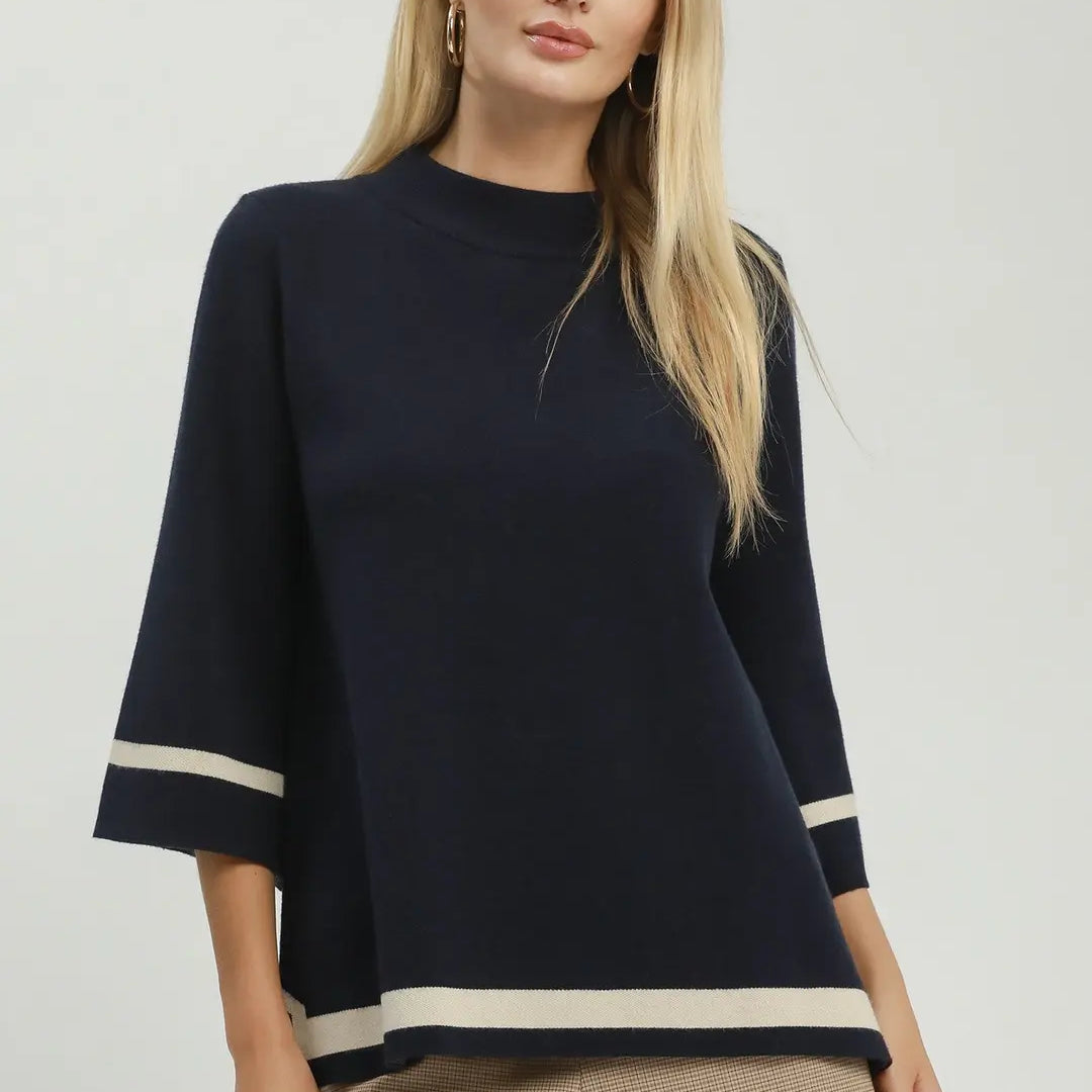A blonde model wearing a navy blue knit top with beige stripes on the sleeves and hem, paired with beige shorts, against a light gray studio background.