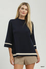 A blonde model wearing a navy blue knit top with beige stripes on the sleeves and hem, paired with beige shorts, against a light gray studio background.