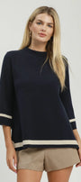 A blonde model wearing a navy blue knit top with beige stripes on the sleeves and hem, paired with beige shorts, against a light gray studio background.