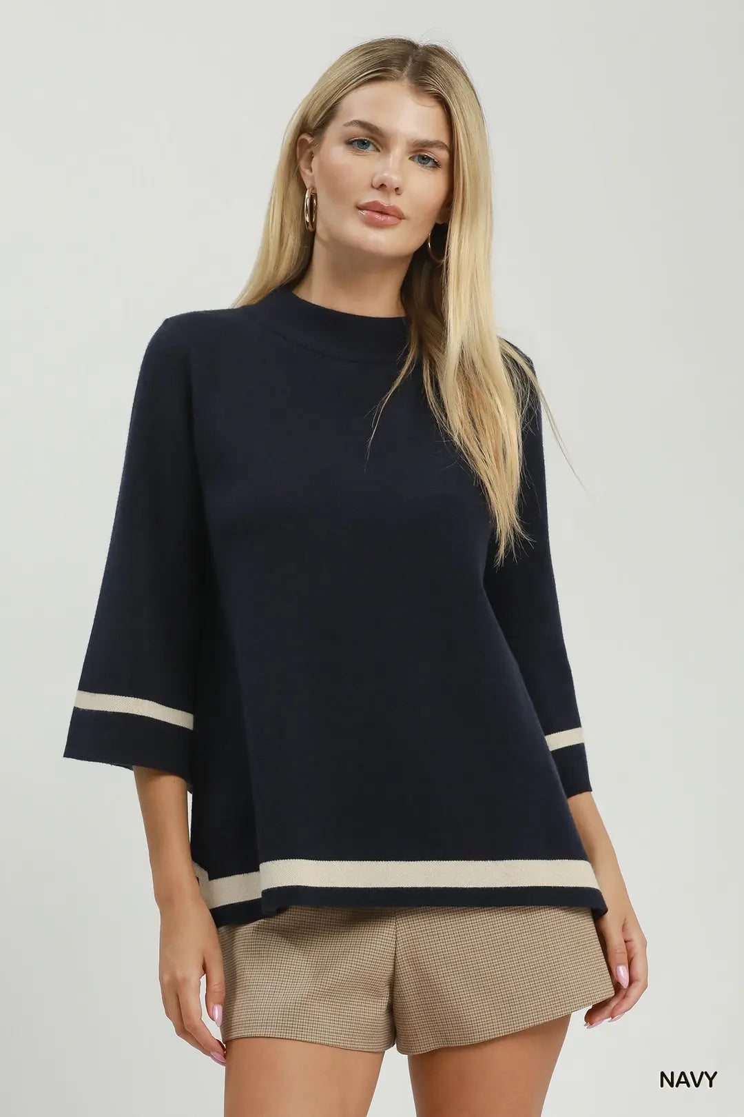 A blonde model wearing a navy blue knit top with beige stripes on the sleeves and hem, paired with beige shorts, against a light gray studio background.