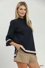 A blonde model wearing a navy blue mock-neck knit top with cream stripes on the sleeves and hem, paired with beige shorts, against a light gray studio background.