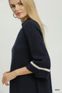 A blonde model wearing a navy blue knit top with three-quarter sleeves and contrasting cream stripes at the cuffs, shown against a light gray studio background.