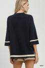 A woman shown from the back wearing a navy blue knit top with cream stripe accents at the sleeve cuffs and hem, paired with beige shorts against a light gray studio background.