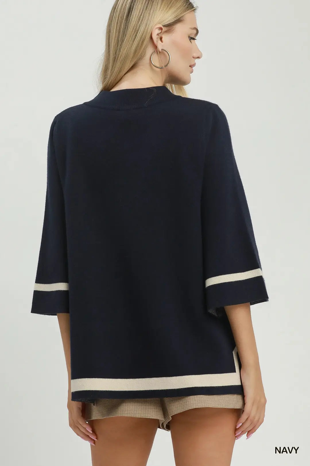 A woman shown from the back wearing a navy blue knit top with cream stripe accents at the sleeve cuffs and hem, paired with beige shorts against a light gray studio background.