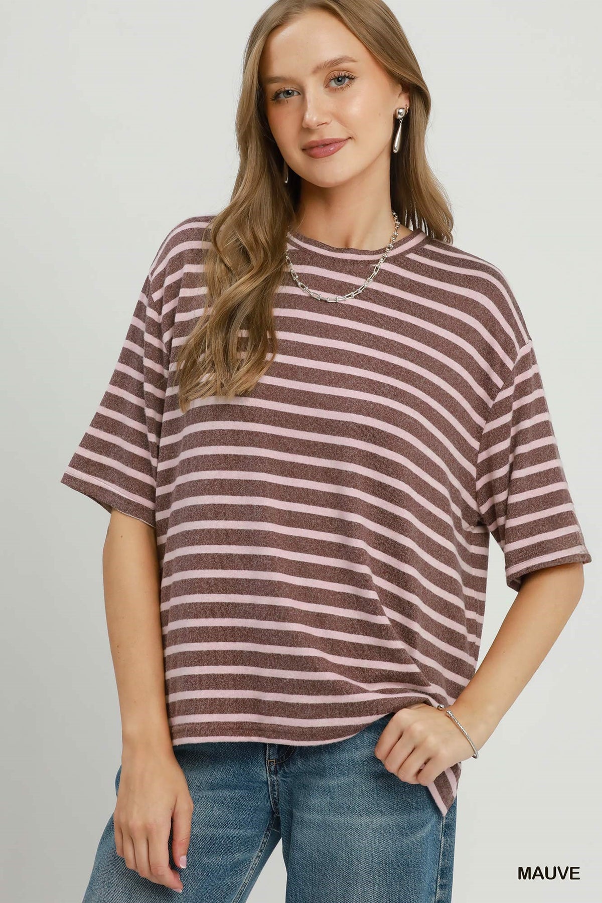 A woman wearing an oversized short-sleeve t-shirt with horizontal mauve and white stripes, paired with blue jeans, against a light gray studio background.
