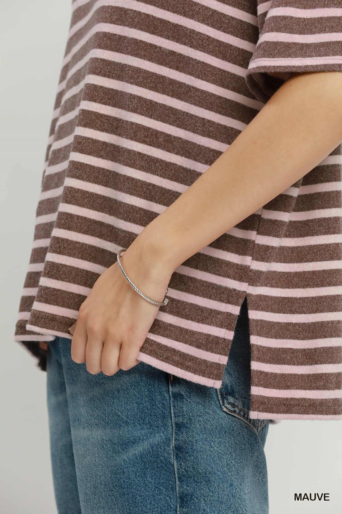 A close-up of a person wearing a brown and pink striped short-sleeve top with blue jeans, showing a thin silver chain bracelet on the wrist. The word 'MAUVE' appears in the bottom right corner.