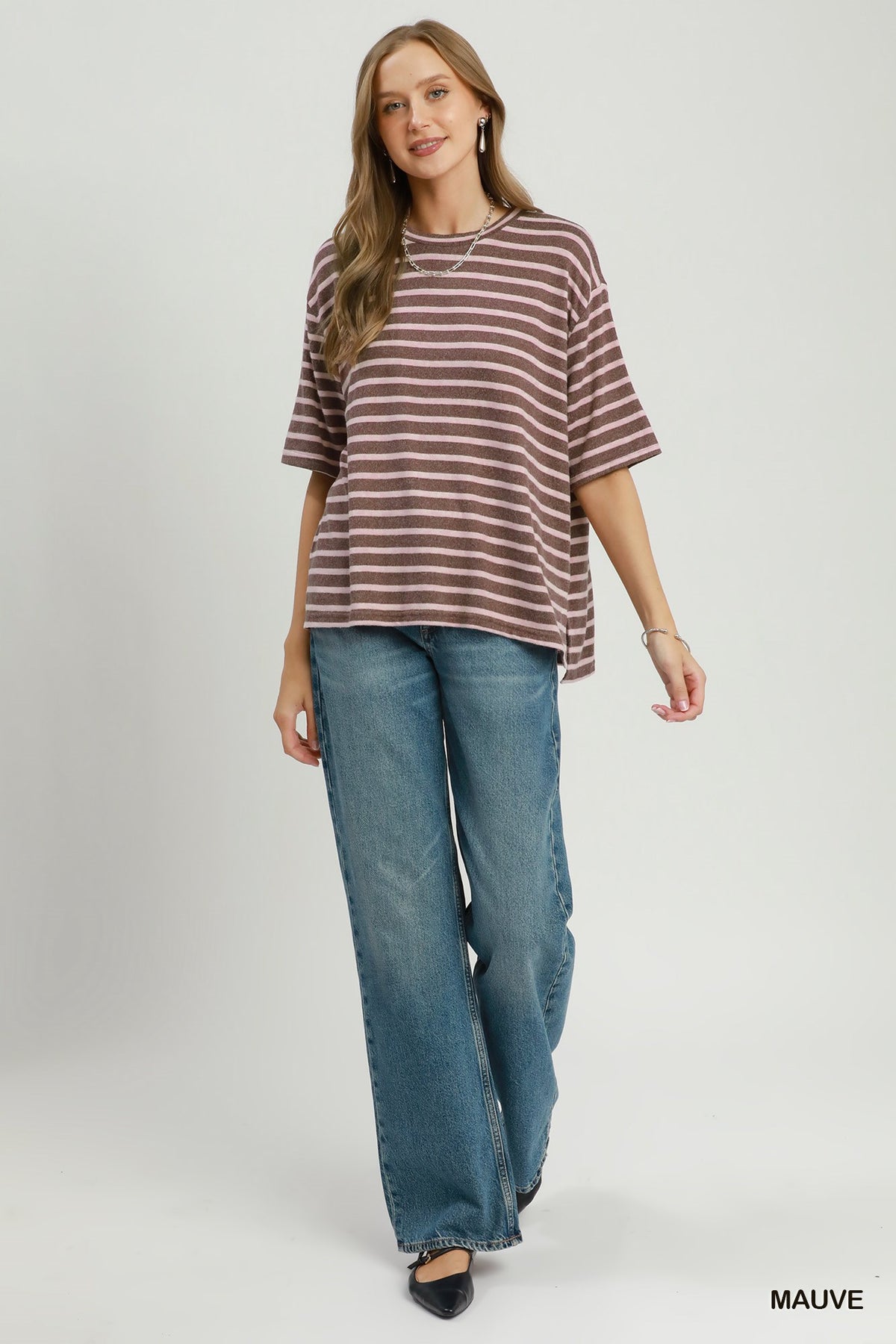 A woman modeling an oversized short-sleeve t-shirt with horizontal mauve and white stripes, paired with wide-leg blue jeans, against a light gray studio background.