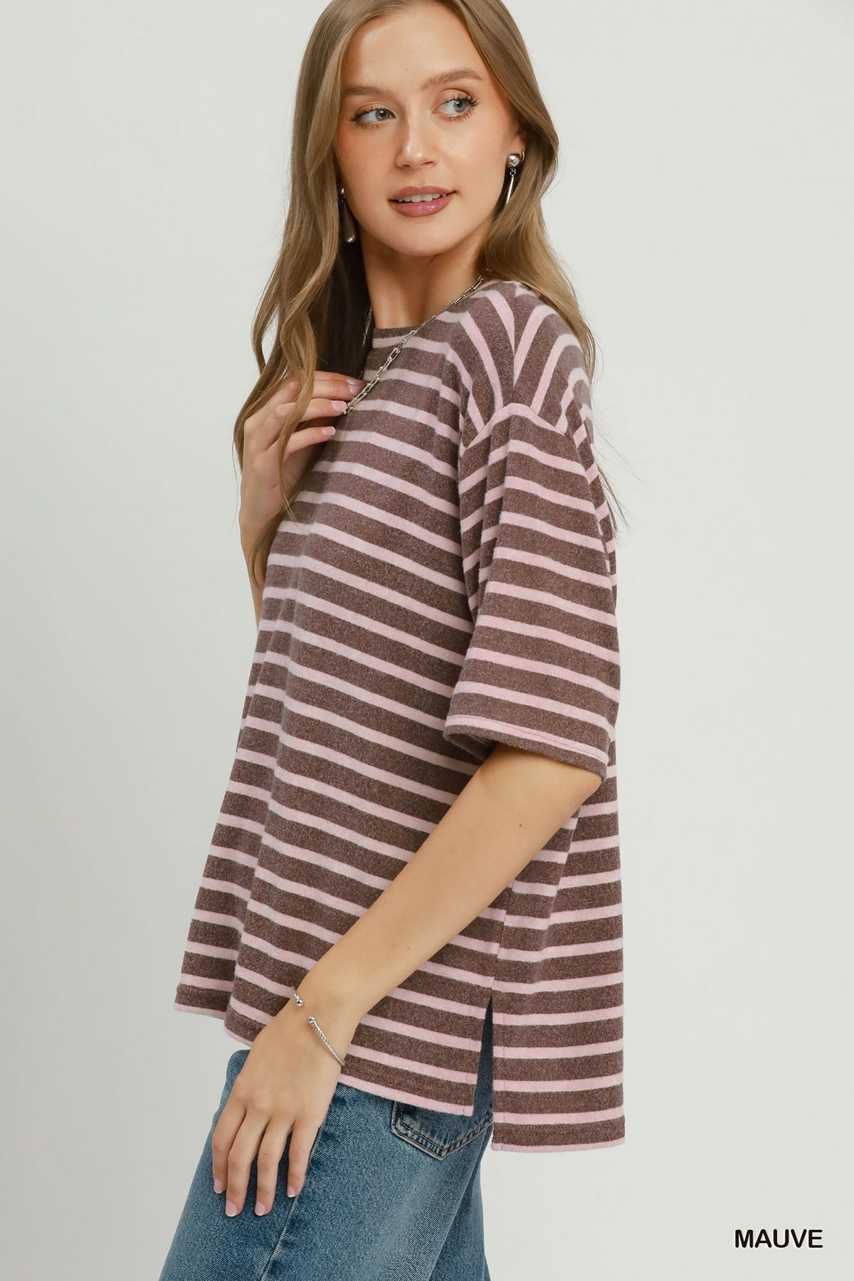 A woman modeling a short-sleeve knit top with horizontal mauve and brown stripes, paired with blue jeans, against a light gray studio background.