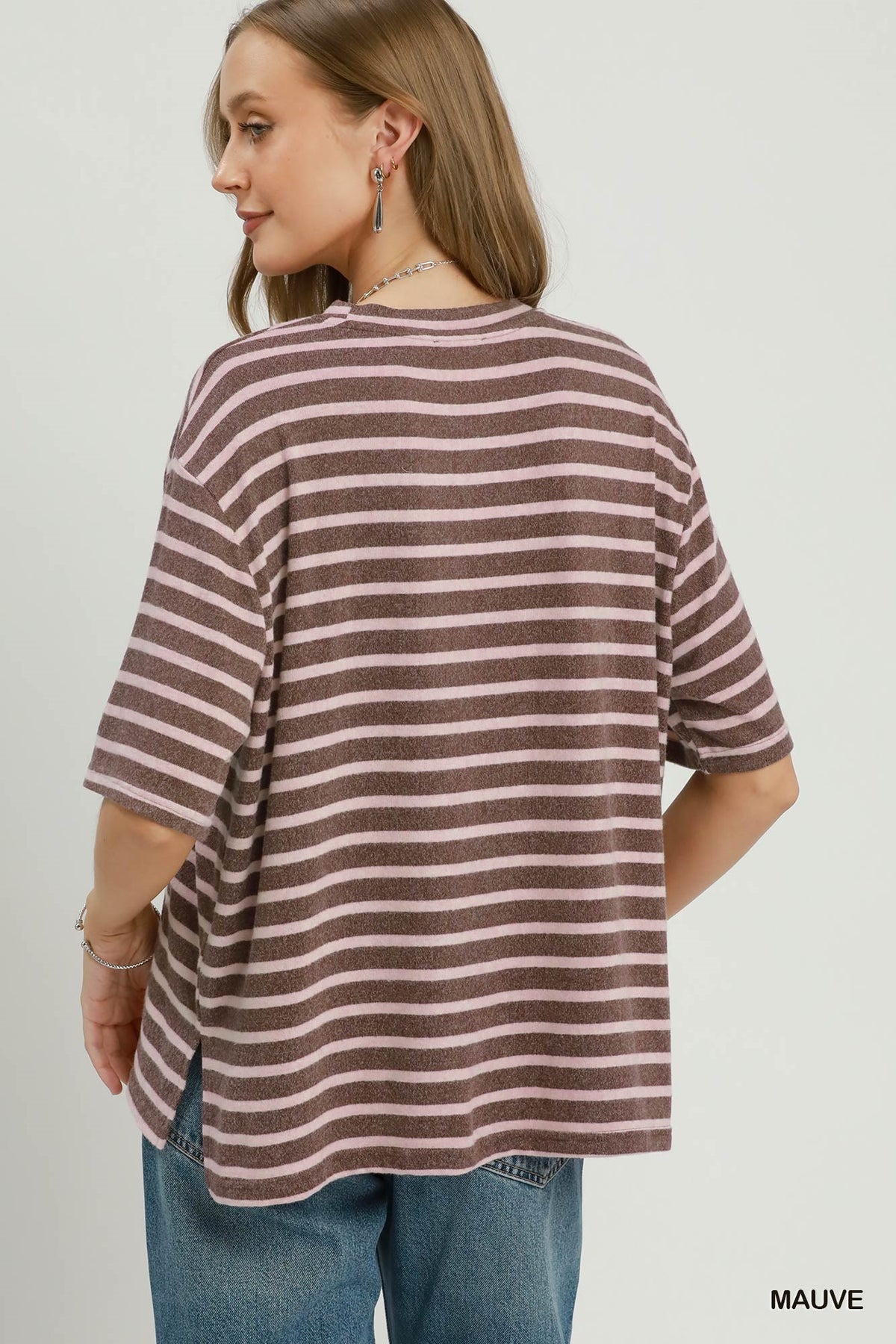 A woman shown from the back wearing an oversized short-sleeve striped top in mauve and off-white, paired with blue jeans, against a plain light gray studio background.