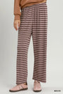 A person wearing loose, wide-leg striped lounge pants in mauve and brown, paired with a tan sweatshirt and pink sneakers, against a plain light gray studio background.