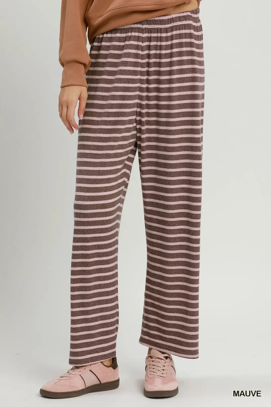 A person wearing loose, wide-leg striped lounge pants in mauve and brown, paired with a tan sweatshirt and pink sneakers, against a plain light gray studio background.