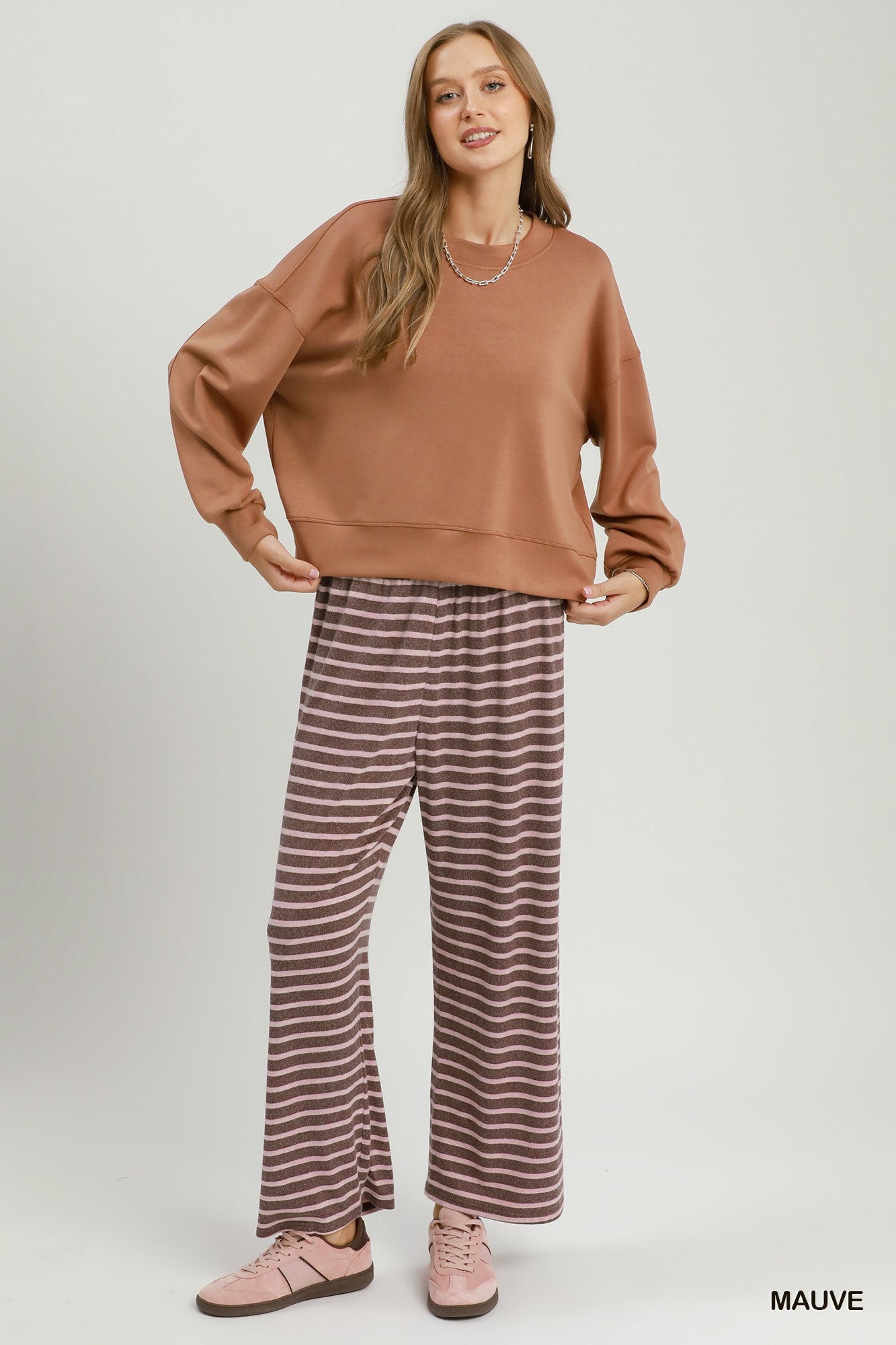 A woman modeling a two-piece lounge set consisting of a cropped mauve sweatshirt and matching wide-leg pants with thin brown stripes, paired with pink sneakers, on a light gray studio background.