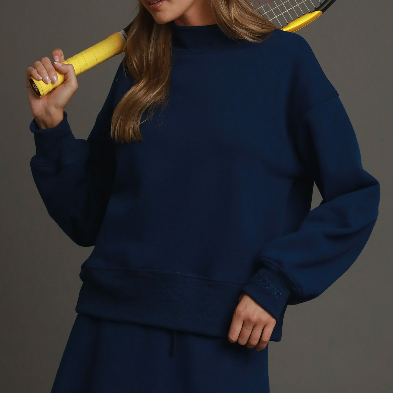 A woman wearing a navy blue sweatshirt and matching mini skirt set, holding a yellow tennis racket over her shoulder, with a white baseball cap. The background is a solid dark gray studio backdrop.