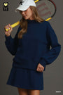 A woman wearing a navy blue sweatshirt and matching mini skirt set, holding a yellow tennis racket over her shoulder, with a white baseball cap. The background is a solid dark gray studio backdrop.