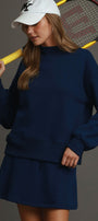 A woman wearing a navy blue sweatshirt and matching mini skirt set, holding a yellow tennis racket over her shoulder, with a white baseball cap. The background is a solid dark gray studio backdrop.