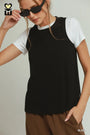 A woman wearing a black sleeveless top over a white t-shirt and brown pants, with black sunglasses, against a light gray background. A small 'BH' logo is visible in the top-left corner, and 'BLACK' is printed in the bottom-right corner.