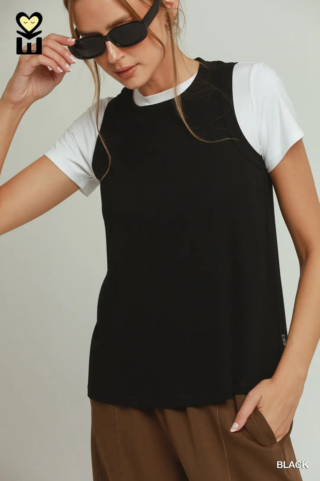 A woman wearing a black sleeveless top over a white t-shirt and brown pants, with black sunglasses, against a light gray background. A small 'BH' logo is visible in the top-left corner, and 'BLACK' is printed in the bottom-right corner.