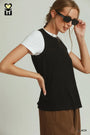 A woman wearing a black sleeveless t-shirt over a white crewneck tee, paired with brown pants and black sunglasses, against a light gray background.