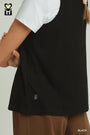 A close-up of a person wearing a black sleeveless tank top over a white t-shirt and brown pants, with a small white embroidered logo near the lower left hem. The brand logo in the top-left corner shows a heart and the letters 'MH'.