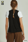 A woman shown from the back wearing a black sleeveless vest over a white short-sleeve t-shirt and brown pants, against a light gray studio background.