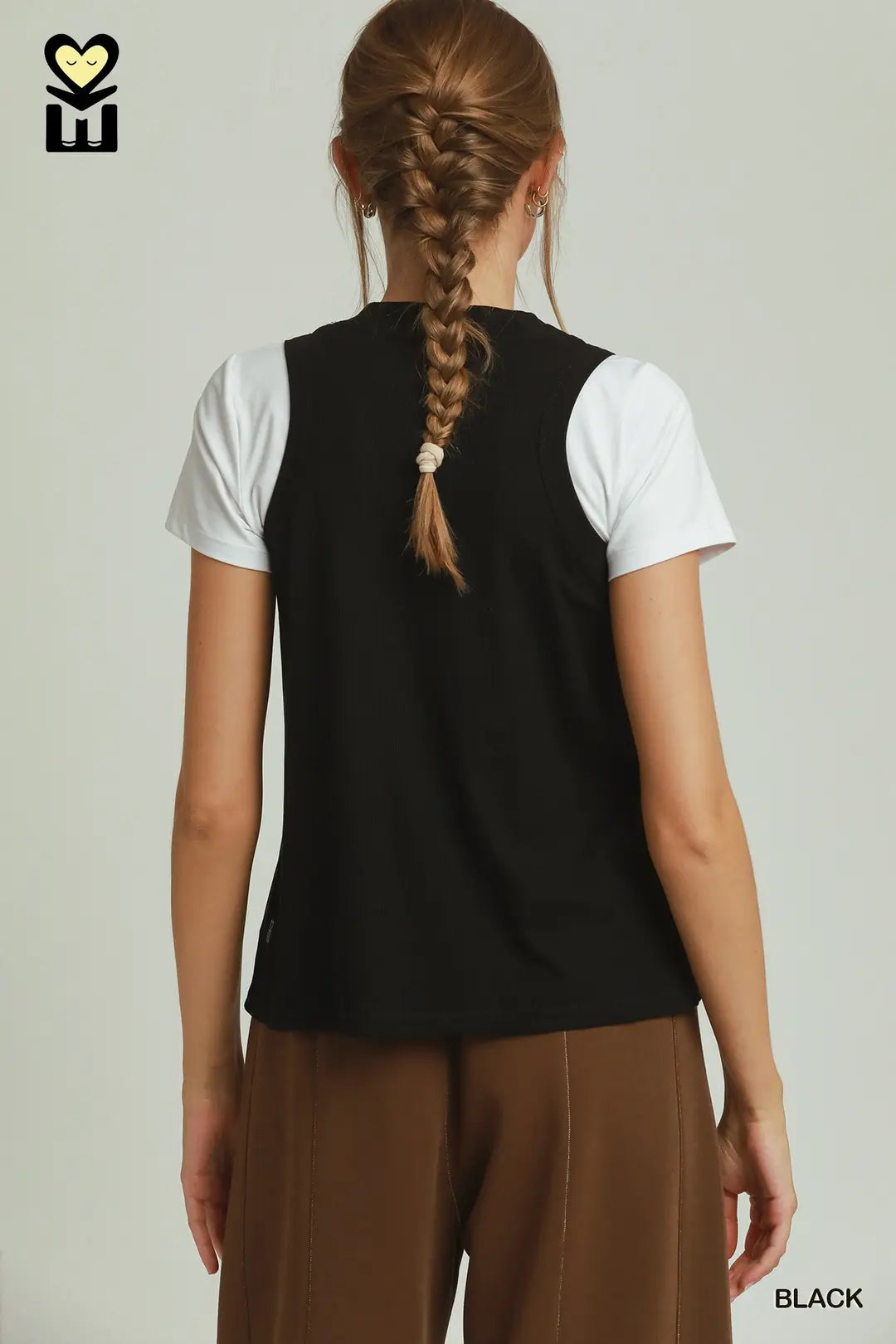A woman shown from the back wearing a black sleeveless vest over a white short-sleeve t-shirt and brown pants, against a light gray studio background.