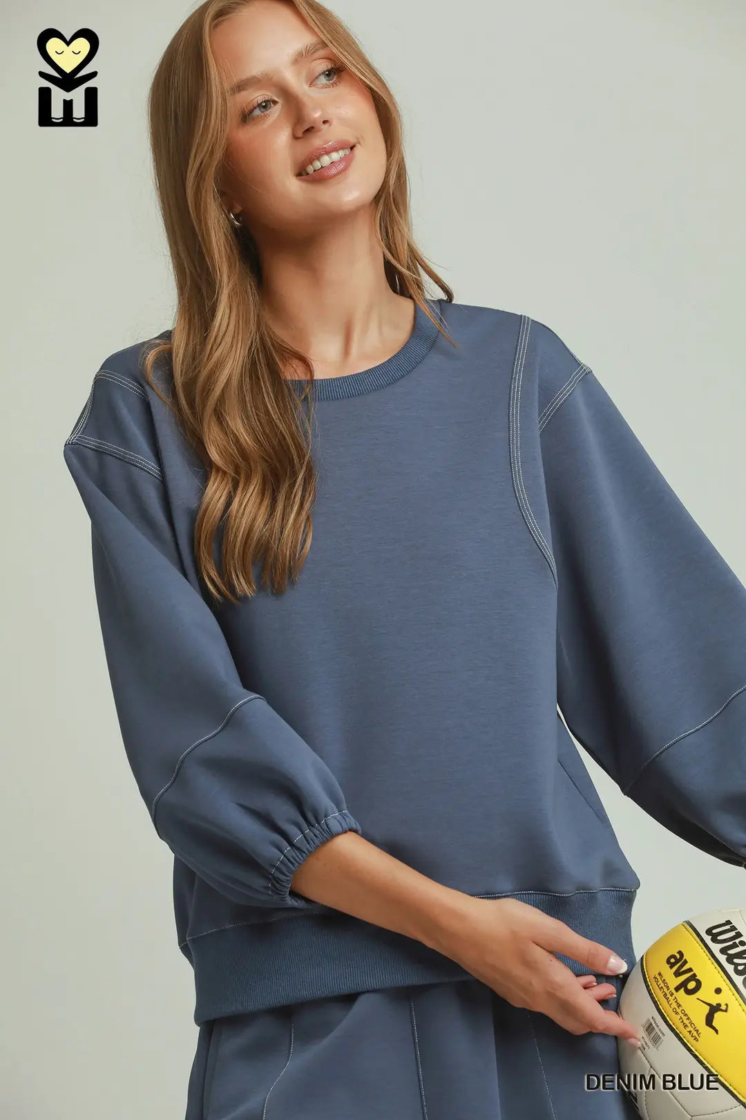 A woman wearing a dusty blue crewneck sweatshirt with three-quarter puff sleeves and elastic cuffs, paired with matching sweatpants. She is holding a yellow and black branded bag against a light gray background.