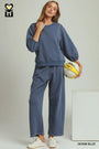 A woman modeling a matching two-piece lounge set in denim blue, featuring a long-sleeve sweatshirt and wide-leg pants, holding a yellow basketball against a light gray studio background.