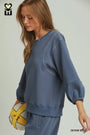A woman wearing a dusty blue sweatshirt and matching sweatpants, holding a yellow and white volleyball. The image features a logo in the top-left corner and the text 'DENIM BLUE' in the bottom-right corner.