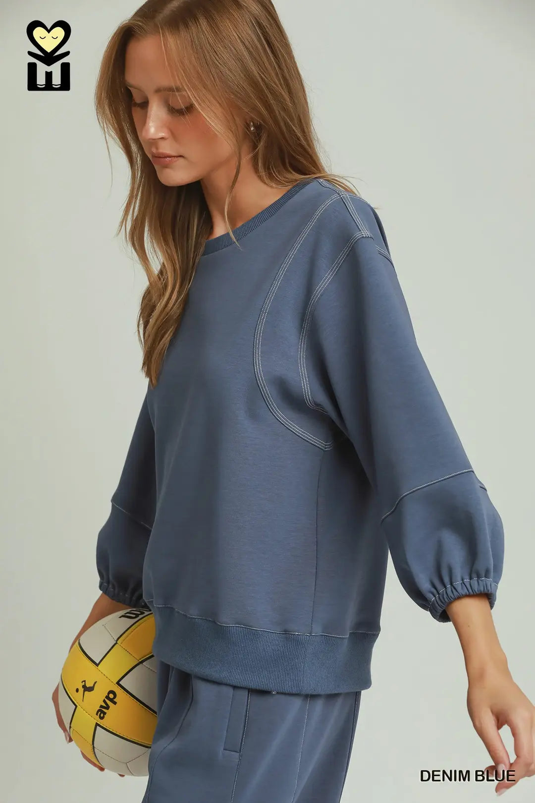 A woman wearing a dusty blue sweatshirt and matching sweatpants, holding a yellow and white volleyball. The image features a logo in the top-left corner and the text 'DENIM BLUE' in the bottom-right corner.