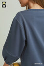 Close-up of a person wearing a loose-fitting blue sweatshirt, showing the back and shoulder area. The sweatshirt is a medium denim blue color with visible seam details.
