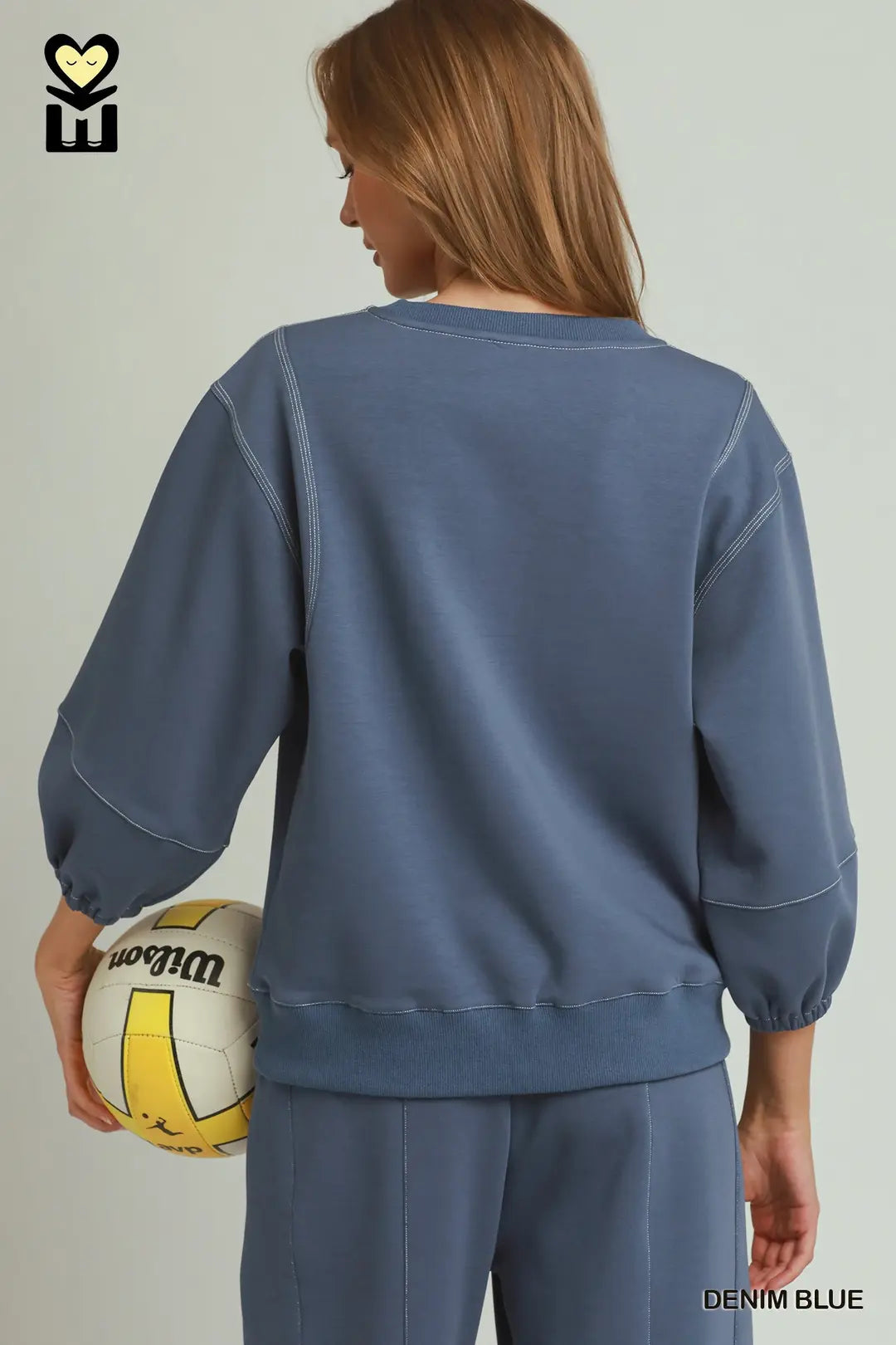 A woman shown from the back wearing a dusty blue sweatshirt and matching sweatpants, holding a yellow and white volleyball. The text 'DENIM BLUE' is visible in the bottom right corner.