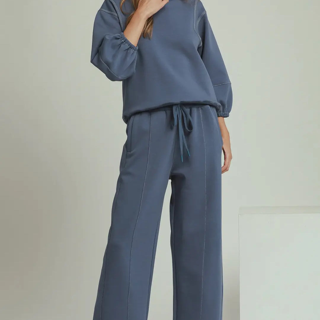 A woman modeling a matching two-piece lounge set in denim blue, featuring a long-sleeve sweatshirt and wide-leg pants with a drawstring waist, paired with white sneakers against a light gray studio background.