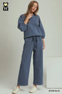 A woman modeling a matching two-piece lounge set in denim blue, featuring a long-sleeve sweatshirt and wide-leg pants with a drawstring waist, paired with white sneakers against a light gray studio background.