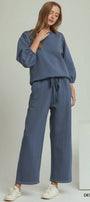 A woman modeling a matching two-piece lounge set in denim blue, featuring a long-sleeve sweatshirt and wide-leg pants with a drawstring waist, paired with white sneakers against a light gray studio background.
