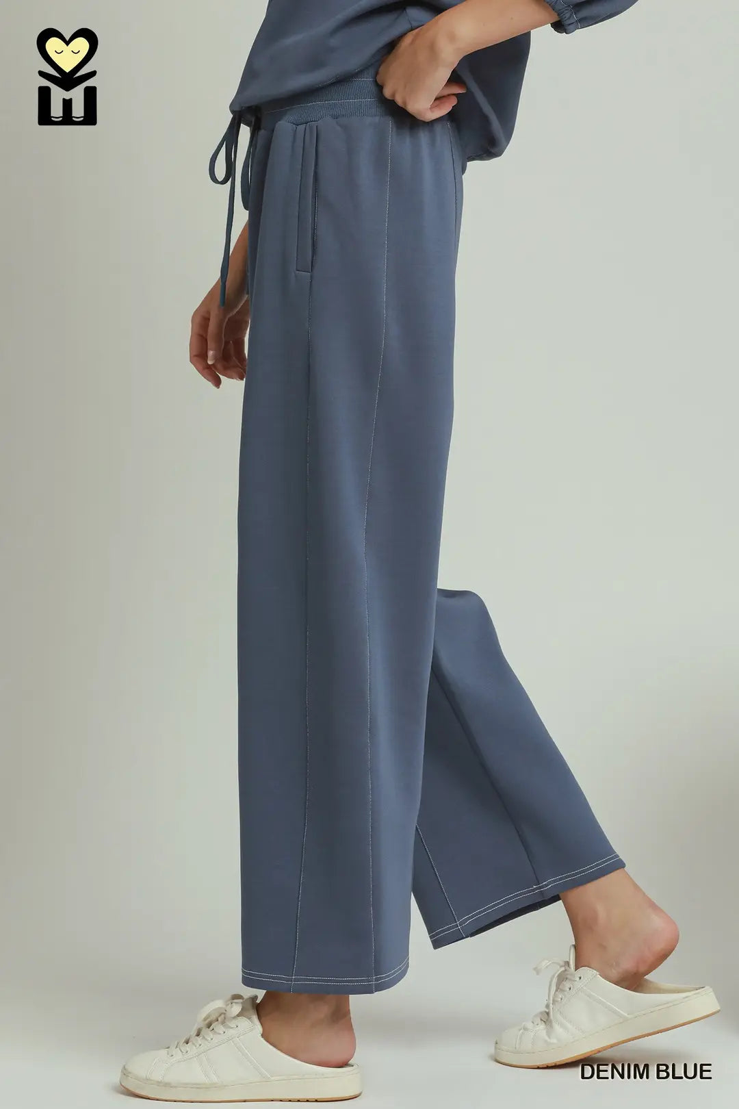 A person wearing wide-leg drawstring pants in denim blue, paired with a matching top and white slip-on sneakers, shown against a plain light gray studio background.