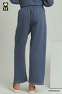 A person shown from the back wearing wide-leg denim blue pants and a matching top, standing indoors against a light neutral wall. The image includes a small logo in the top-left corner and the text 'DENIM BLUE' in the bottom-right corner.