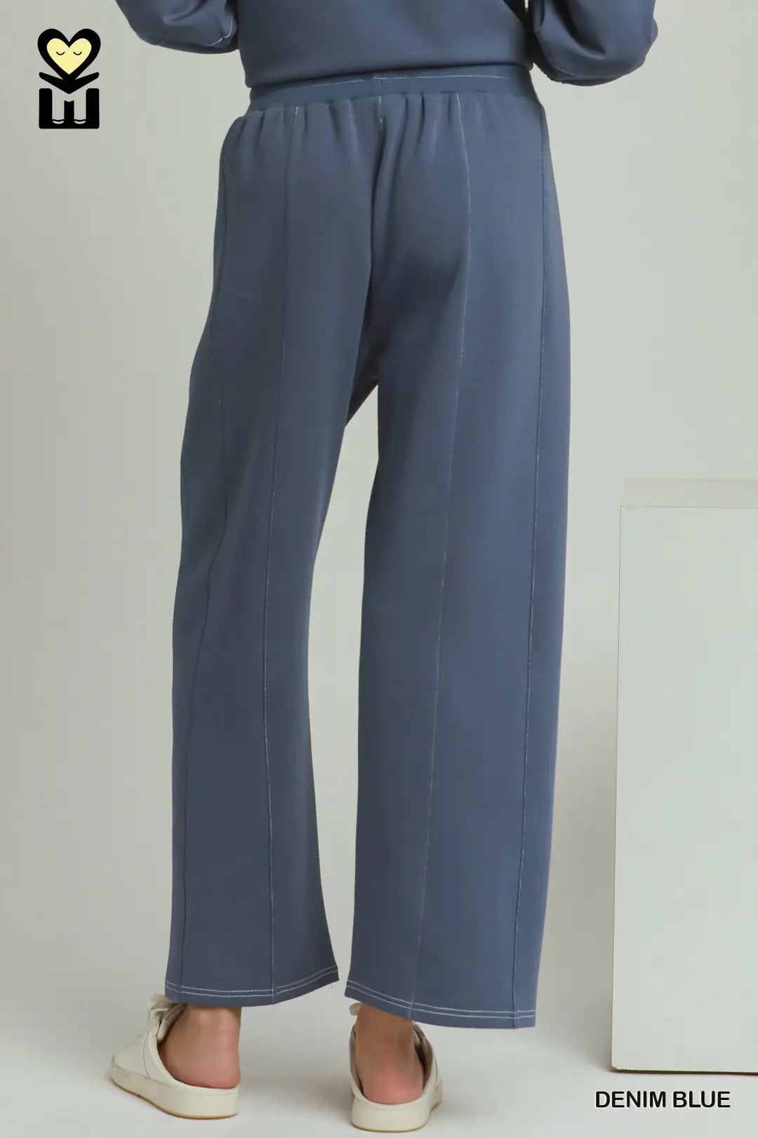 A person shown from the back wearing wide-leg denim blue pants and a matching top, standing indoors against a light neutral wall. The image includes a small logo in the top-left corner and the text 'DENIM BLUE' in the bottom-right corner.