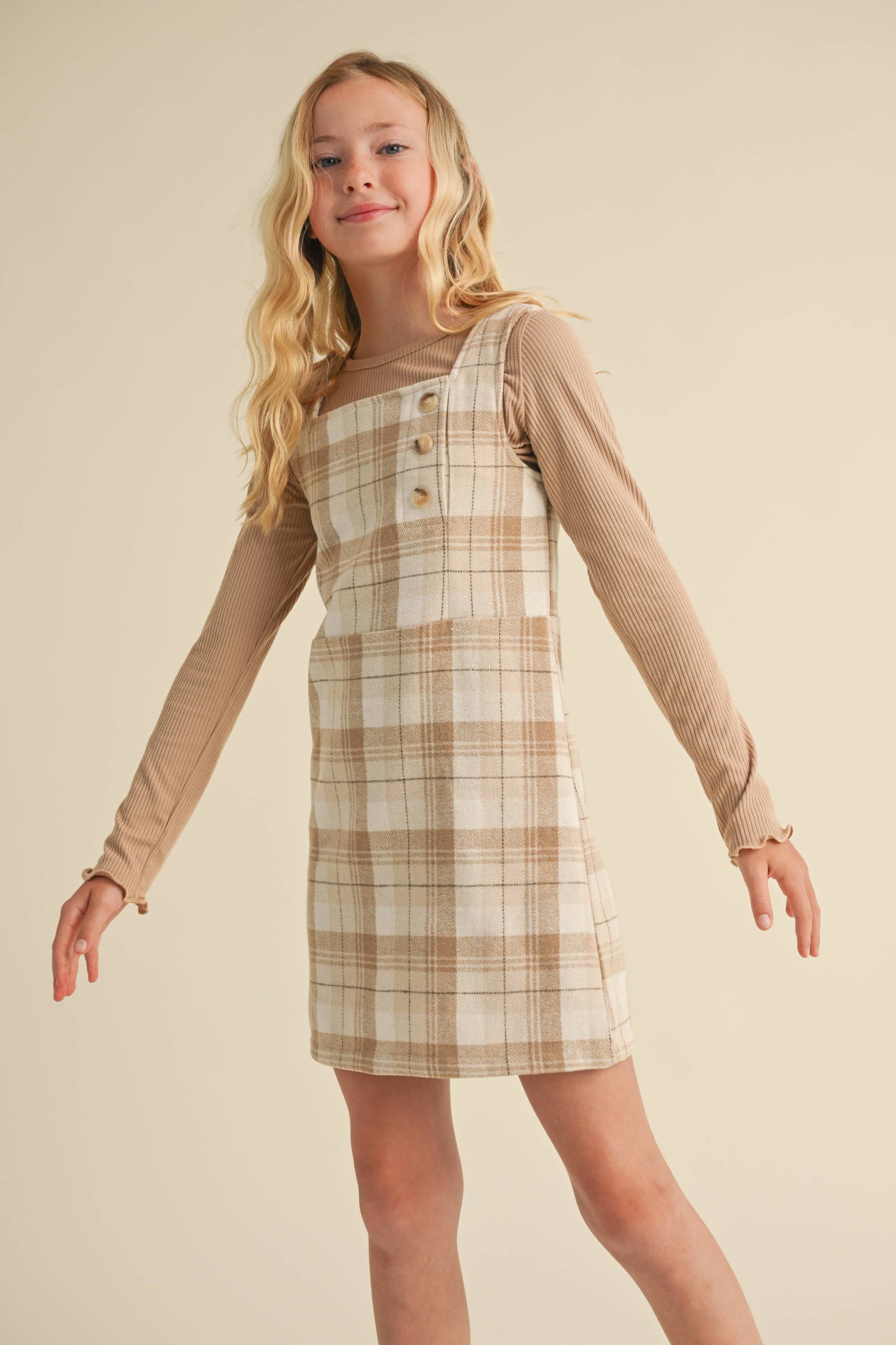 Young girl wearing a plaid dress over a long-sleeve shirt against a beige background