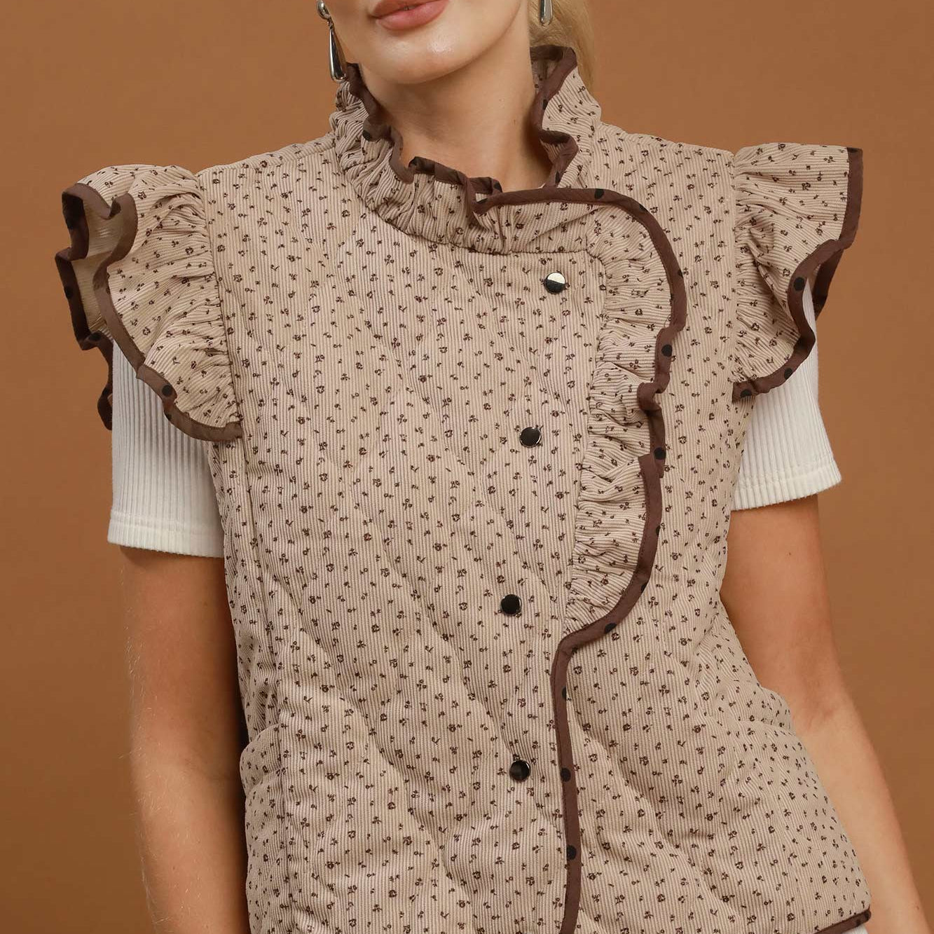 A woman models a sleeveless quilted vest with ruffled trim and a small-scale floral pattern in beige and brown, worn over a white short-sleeve top and light blue jeans against a solid brown studio background.