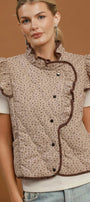 A woman models a sleeveless quilted vest with ruffled trim and a small-scale floral pattern in beige and brown, worn over a white short-sleeve top and light blue jeans against a solid brown studio background.