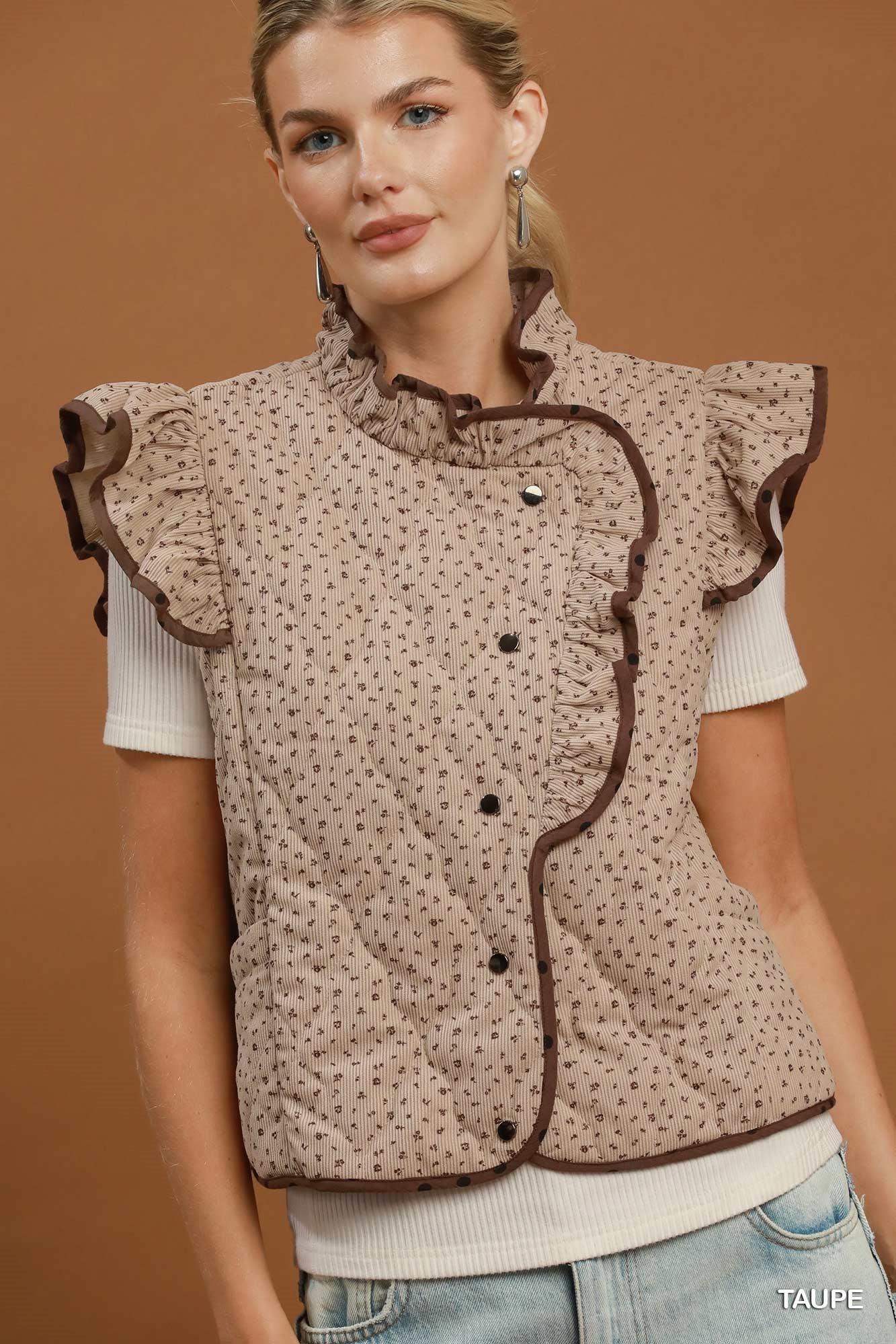 A woman models a sleeveless quilted vest with ruffled trim and a small-scale floral pattern in beige and brown, worn over a white short-sleeve top and light blue jeans against a solid brown studio background.