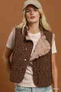 A woman wearing a taupe brown quilted vest with black polka dots and a ruffled light beige collar, styled over a cream ribbed t-shirt and a beige cap against a solid brown background.