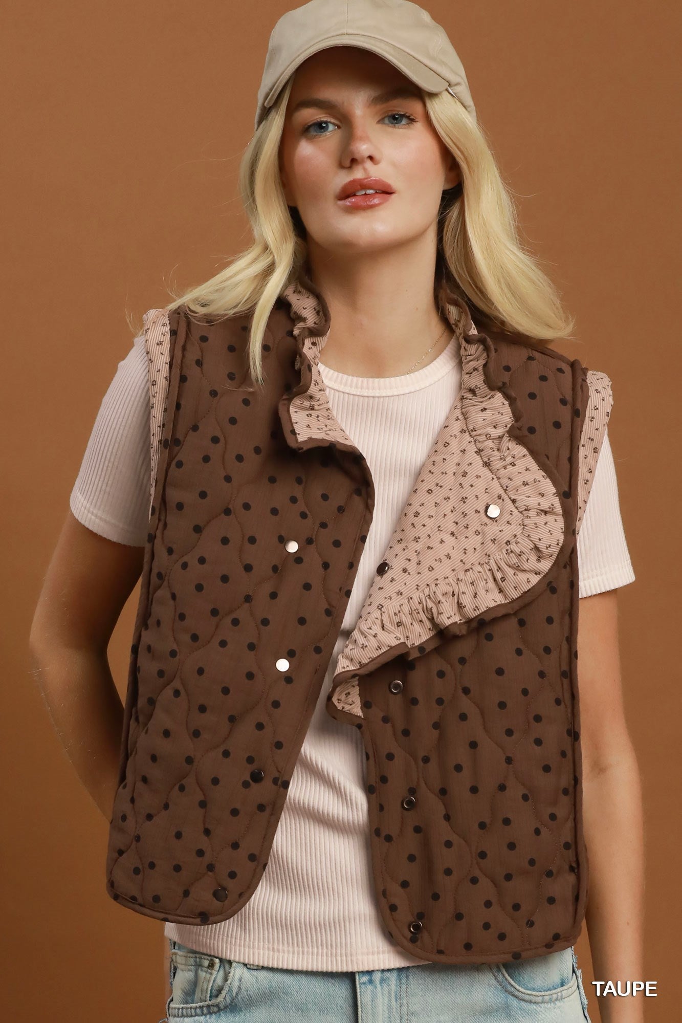 A woman wearing a taupe brown quilted vest with black polka dots and a ruffled light beige collar, styled over a cream ribbed t-shirt and a beige cap against a solid brown background.